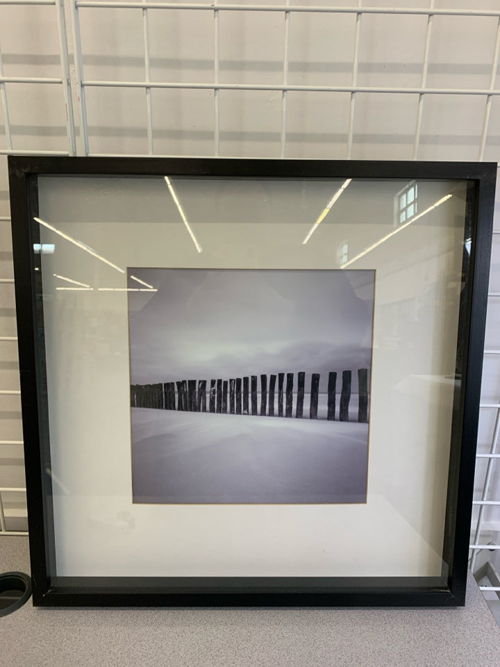 WOOD PILLARS ON BEACH PHOTO IN BLACK FRAME.