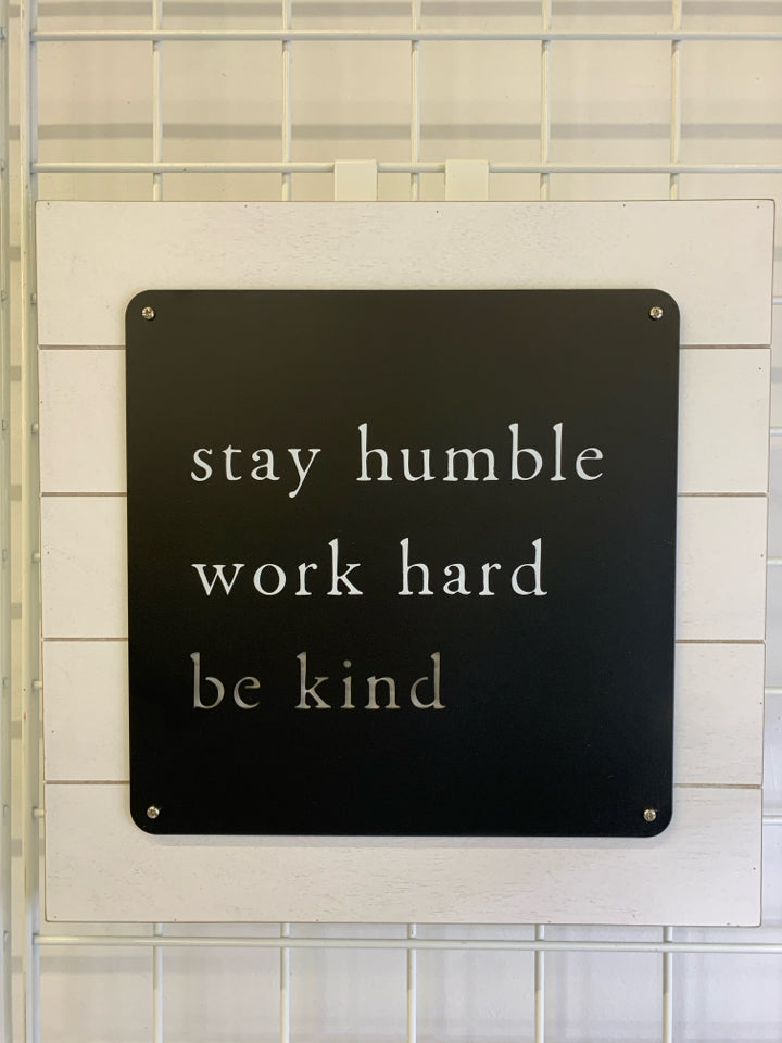 STAY HUMBLE WORK HARD KIND METAL CUT-OUT SIGN ON WHITE WOOD.